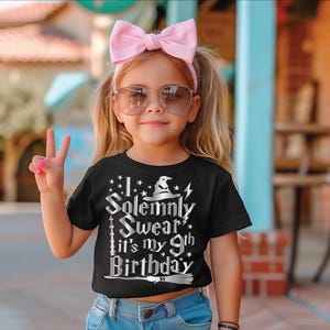 May include: A young girl wearing a black t-shirt with the text "I Solemnly Swear it's my 9th Birthday" in silver lettering. She is wearing a pink bow headband and sunglasses. The girl is smiling and giving a peace sign.