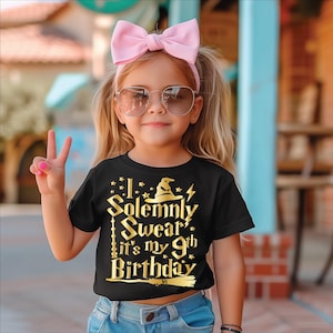 May include: A young girl wearing a black t-shirt with gold text that reads "I Solemnly Swear it's my 9th Birthday". She is wearing a pink bow headband and sunglasses. The girl is holding up two fingers in a peace sign.