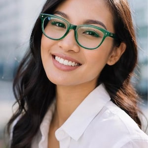 May include: A smiling person wearing green-framed eyeglasses. The glasses have a classic shape and complement the person's dark hair and white collared shirt. The background is blurred.