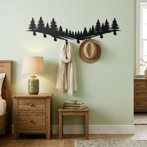 May include: A black metal wall-mounted coat rack shaped like a forest silhouette. A beige scarf and a straw hat hang from the rack. The rack is mounted on a light green wall, with a wooden nightstand and a lamp to the left.