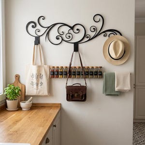 May include: A black metal wall-mounted spice rack with a decorative scroll design. A beige tote bag, a brown leather satchel, a straw hat, and two towels hang from the rack. A row of spice jars is mounted below the decorative scroll.