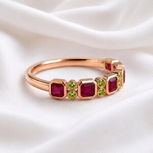 May include: A rose gold ring featuring a series of alternating square-cut deep red rubies and pairs of small round-cut bright green peridot stones set along the band, creating a vibrant contrast of colors in a polished metal setting.