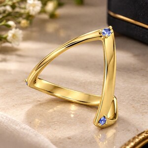 May include: A polished gold ring featuring an open, triangular geometric design. The band is accented with three small, round, blue gemstones set at the corners of the structure, creating a distinct and modern look with a smooth, reflective finish.