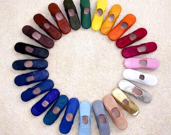 May include: A collection of twenty-four leather slippers arranged in a circular pattern. The slippers feature a variety of colours including black, brown, green, yellow, orange, red, pink, white, silver, gold, beige, grey, and multiple shades of blue.