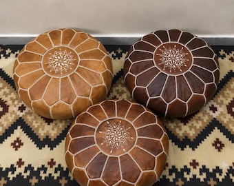 May include: Three round leather poufs in varying shades of tan, medium brown, and dark brown. Each pouf features a central white embroidered star-like pattern and white stitched seams radiating outward in a segmented, circular design.