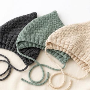 Puede incluir: Tres gorros de bebé tejidos en carbón, verde salvia y crema. Cada gorro tiene una corona redondeada y un borde acanalado, con lazos a juego para asegurar debajo de la barbilla. Los gorros están colocados sobre una superficie blanca.