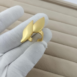 May include: A gold-toned ring featuring two smooth, leaf-shaped elements that extend outward from the central band. The metal has a polished, reflective finish, and the design consists of two distinct, curved, petal-like shapes joined together.