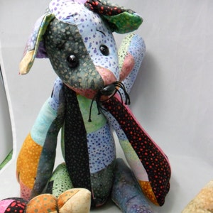 May include: A handmade patchwork dog plush toy with a variety of colorful fabric patterns. The dog has black button eyes, a black nose, and long, floppy ears. The patchwork includes polka dots, floral prints, and solid colors.
