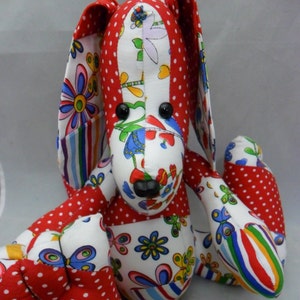 Op de afbeelding: Een handgemaakt patchwork konijn speelgoed van rode, witte en blauwe stof. Het konijn heeft een wit gezicht met zwarte ogen en een zwarte neus. De oren van het konijn zijn lang en slap.