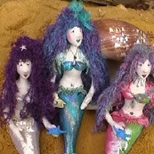 May include: Three handmade mermaid dolls with vibrant, colourful designs. Each doll has a unique hairstyle and a shimmering tail. The mermaids are posed on a sandy surface, with one holding a small blue fish. The dolls are approximately 25 cm tall.