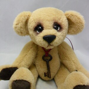 May include: A soft, tan teddy bear with dark brown detailing around the eyes and on the paws. The bear has a black nose and a vintage key pendant suspended from a red ribbon. The bear is seated with its legs outstretched.