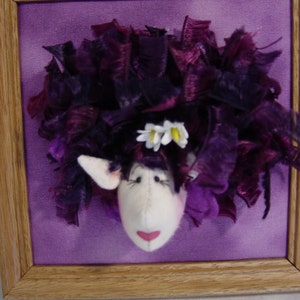May include: A framed textile artwork depicting a sheep's head. The sheep has a white face, pink mouth, and dark purple, ribbon-like hair. It is decorated with small white flowers. The background is a solid purple colour, and the frame is wooden.