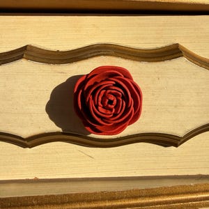 May include: A close-up of a red rose-shaped decorative element on a light-colored wooden surface. The rose is centered, with detailed petals. The wood has a gold trim and a light yellow color.
