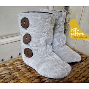 以下が含まれることがあります： 茶色のボタンが付いた灰色のふわふわのスリッパ。スリッパは籐のバスケットの上に置かれています。