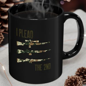 May include: Black ceramic mug featuring the text "I Plead The 2nd" and three rifles with camouflage designs. The mug is set on a light grey surface, with decorative elements in the background.