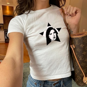 Puede incluir: Camiseta blanca con un gráfico de estrella negra con un retrato en blanco y negro. La camiseta es de manga corta y parece estar hecha de un material suave. La modelo lleva vaqueros azules y un bolso marrón.