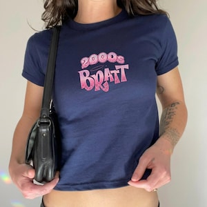 May include: Navy blue crop top t-shirt with the text "2000s Bratt" in pink. The shirt has short sleeves and a round neckline. A black shoulder bag is visible on the left side.