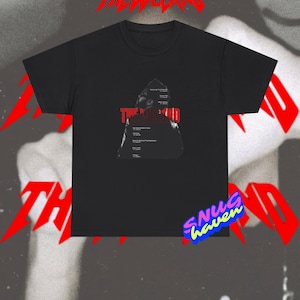 May include: A black short-sleeve t-shirt featuring a graphic print of The Weeknd. The design includes the artist's name in bold red text and a list of album titles in white font. The shirt has a crew neckline and a standard straight hem.
