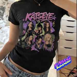 May include: A black short-sleeve t-shirt featuring a graphic print of the word KATSEYE in purple text above six illustrated figures. The design includes colorful heart shapes, stars, and lightning bolt accents in shades of purple, green, and pink.