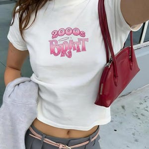 May include: White t-shirt with the text "2000s BRATT" in pink, featuring a halo above the "B". A red handbag with a zipper is visible. The outfit includes a gray hoodie, gray pants, and a pink belt.