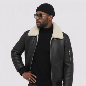 May include: A black bomber jacket with a white faux shearling collar, worn over a black turtleneck sweater. The outfit includes a black ribbed beanie, gold-rimmed aviator sunglasses with gradient lenses, and a gold ring on the finger.