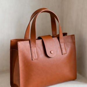 Op de afbeelding: Een bruine leren tote bag met twee handvatten aan de bovenkant en een klepsluiting met een knoop. De tas heeft een rechthoekige vorm en een minimalistisch ontwerp.