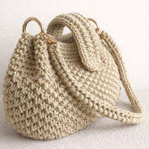 Könnte beinhalten: Beige gehäkelte Handtasche mit runder Form und strukturiertem Griff. Die Tasche hat goldfarbene Metallringe und einen langen, verstellbaren Riemen. Das Häkelmuster ist eng gewebt, was der Tasche ein robustes Aussehen verleiht.