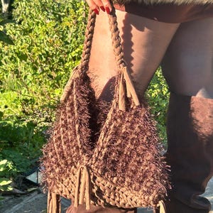May include: A brown crochet handbag with a braided handle and fringe tassels. The bag has a textured, shaggy appearance and is held in front of a green background. The bag is a unique accessory.