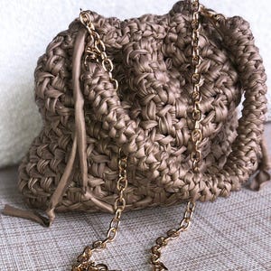 Può includere: Una borsa a mano intrecciata color talpa con una tracolla a catena dorata. La borsa ha una forma arricciata, simile a una nuvola, e una chiusura con coulisse. La catena è attaccata alla borsa con elementi dorati.