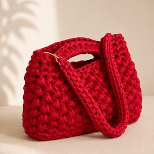 Può includere: Una vivace borsa a mano rossa all'uncinetto, dalla forma strutturata. La borsa presenta un manico superiore e una tracolla staccabile, entrambe realizzate con lo stesso filato spesso e strutturato. Una chiusura color oro assicura la tracolla. La borsa è su una superficie neutra.