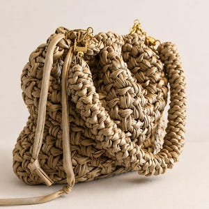 May include: A beige woven handbag with a drawstring closure and a braided handle. The bag features a textured, basket-weave design and gold-colored hardware. The bag has a shoulder strap and is made of a soft, flexible material.