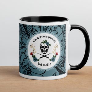 Könnte beinhalten: Weißer Keramikbecher mit schwarzem Innenraum und Henkel. Der Becher hat ein blaugrünes Blumenmuster und ein rundes Design mit einem Totenkopf, gekreuzten Knochen und dem Text "the horrors persist but so do I."
