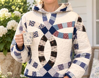 May include: A long-sleeve hooded pullover constructed from quilted fabric featuring a double wedding ring pattern. The garment displays a cream-colored base with intricate patchwork sections in shades of blue, black, and white floral and geometric prints.