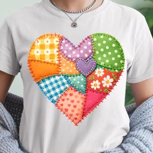 Puede incluir: Camiseta blanca con un diseño de corazón de patchwork colorido. El corazón está compuesto por varios patrones de tela, incluyendo lunares, cuadros y estampados florales, en tonos naranja, verde, rosa y azul. Un pequeño corazón morado está en el centro.