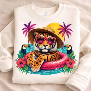 Puede incluir: Sudadera color crema con un tigre con gafas de sol y un sombrero de paja, flotando en una boya rosa. El diseño incluye tucanes, palmeras y flores de hibisco, con un sol amarillo en el fondo.