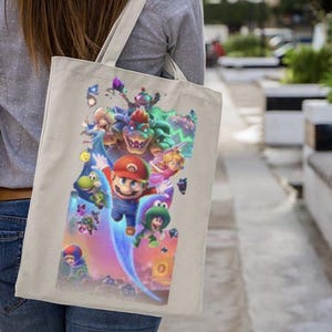 Puede incluir: Bolso tote beige con un gr&aacute;fico colorido de Mario y otros personajes de la pel&iacute;cula Super Mario Bros. La imagen incluye a Mario, la Princesa Peach, Yoshi y Bowser. El bolso tiene un dise&ntilde;o sencillo con una correa para el hombro.