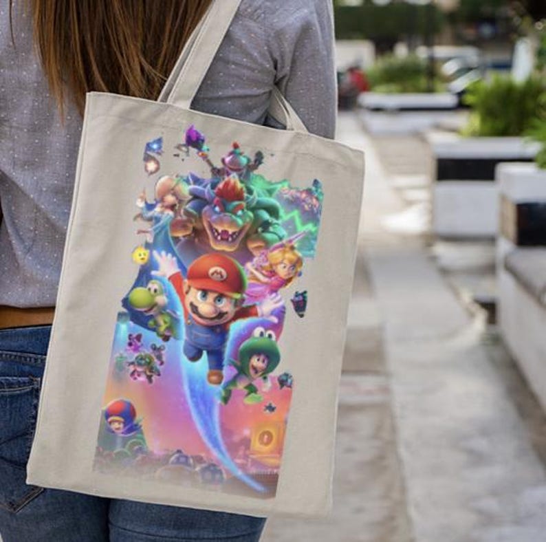 Puede incluir: Bolso tote beige con un gr&aacute;fico colorido de Mario y otros personajes de la pel&iacute;cula Super Mario Bros. La imagen incluye a Mario, la Princesa Peach, Yoshi y Bowser. El bolso tiene un dise&ntilde;o sencillo con una correa para el hombro.