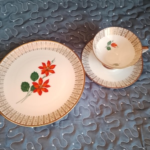 Könnte beinhalten: Ein Vintage-Teeservice mit einem Teller, einer Untertasse und einer Teetasse. Die weißen Porzellanstücke haben einen Goldrand und ein dekoratives Muster. Ein roter Weihnachtsstern mit grünen Blättern ist auf Teller und Tasse gemalt.