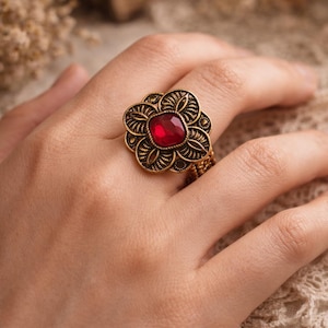 Peut inclure: Bague dorée ornée d'un motif floral détaillé. Elle présente une pierre rouge foncé, taillée en carré, au centre. La bague, portée au doigt, met en valeur son esthétique ornée et d'inspiration vintage.