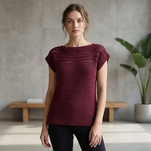 May include: A burgundy crochet top with a boat neck and short sleeves. The top has a textured pattern and is worn with black leggings. A wooden bench and potted plant are in the background.