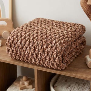 May include: A folded, plush, brown blanket with a textured, woven design. The blanket is made of a soft material and is folded neatly on a wooden shelf. The blanket is a warm, neutral color.