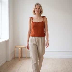 May include: A woman in a rust-coloured, textured knit vest top with tie straps and beige linen trousers. The top has a square neckline. The outfit is casual and comfortable. The background is a white wall and a light wood floor.