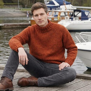 May include: A rust-colored, knitted sweater with a high neckline. The sweater is paired with dark gray jeans and brown leather boots. The person is sitting on a wooden dock near boats.