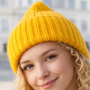 Puede incluir: Primer plano de una persona que lleva un gorro de punto amarillo brillante. El gorro es acanalado y tiene un borde doblado. La persona tiene la piel clara, el pelo rubio rizado y está sonriendo.