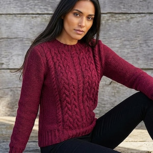 May include: A burgundy cable-knit jumper with a crew neck. The jumper features a textured cable design on the front and sleeves. The model is wearing black trousers. The background is a weathered wooden wall.