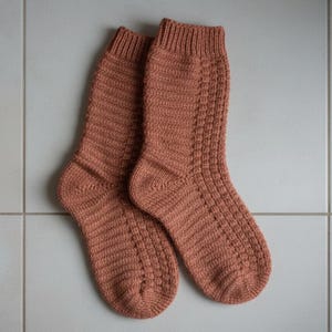 May include: A pair of knitted socks in a warm, earthy orange-brown hue. The socks feature a ribbed cuff and a textured pattern on the leg and foot. The socks are made of a soft, possibly wool, material.