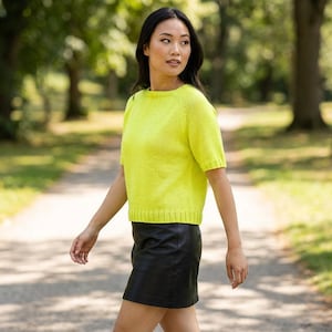 May include: A woman wearing a short-sleeved, bright yellow knit sweater and a black leather skirt. The sweater has a crew neck and ribbed hem. The woman is walking on a paved path in a park with trees in the background.