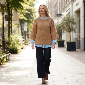Könnte beinhalten: Eine Frau trägt einen beigefarbenen Häkelpullover mit weiten Ärmeln, ein hellblaues Hemd und dunkelblaue Hosen. Sie geht auf einem gepflasterten Bürgersteig, gesäumt von Bäumen und Gebäuden. Der Pullover hat eine warme, neutrale Farbe.