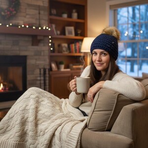 May include: A woman wearing a navy blue knitted hat with a faux fur pom-pom, holding a mug. She is sitting on a beige sofa, wrapped in a cream cable knit blanket. A fireplace with a burning fire is in the background.
