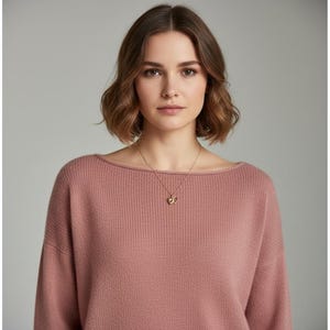 Pode incluir: Uma mulher vestindo um suéter rosa com decote canoa. Um colar dourado com um pingente é visível. O fundo é cinza neutro sólido.